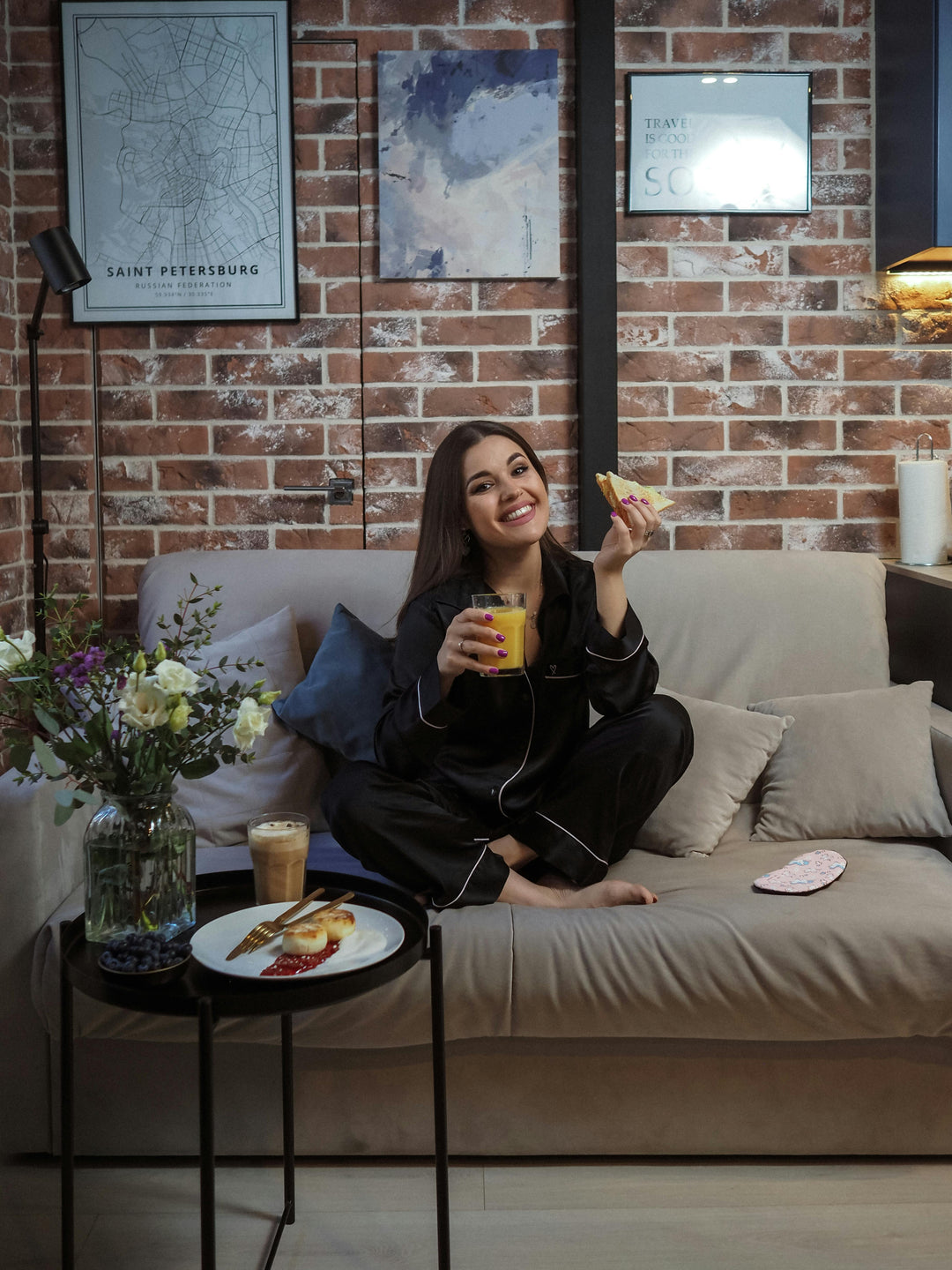 Une femme souriante sur un canapé avec pyjama en satin noir, une tasse de thé à la main